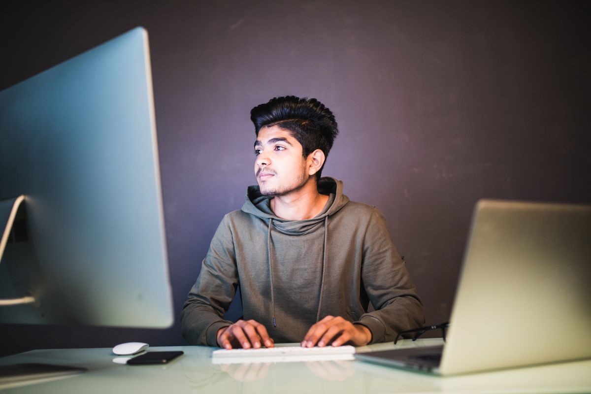 Young Man Sitting At Desk Working On Computer And Laptop At Home